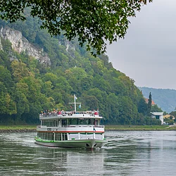 Fahrgastschiff auf Fluss vor bewaldetem Hang und Dorf mit Kirchturm bei bewölktem Himmel